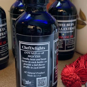 May include: A blue glass bottle with a black label that reads "ChefDelights" and "INFUSED 12-18 MONTHS". The label also includes the text "Vanilla bean and bean caviar in each bottle; we provide a full flavor profile in each bottle". The bottle has a black cap and a QR code on the label. The bottle is sitting on a table with a red tassel.