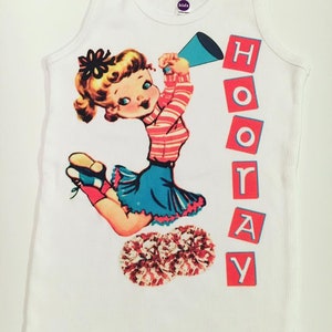 May include: White tank top with a vintage illustration of a cheerleader holding a megaphone and pom-poms. The text "HOORAY" is printed in red blocks on the right side of the image.