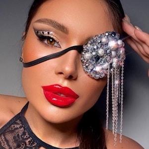 May include: A woman wearing a black eye patch decorated with rhinestones and a silver chain. She has red lipstick and is wearing a black lace top.