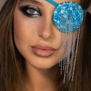May include: A circular eye patch featuring a dense arrangement of light blue, turquoise, and white beads. The patch is attached to a thin turquoise strap and includes several long, dangling silver-toned metal chains that hang down from the bottom edge.