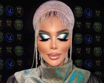 May include: A person wearing a sparkling headpiece with dangling crystal strands. The makeup features bold blue and white eyeshadow, and full, glossy lips. The person is also wearing a metallic scarf with blue and green accents.