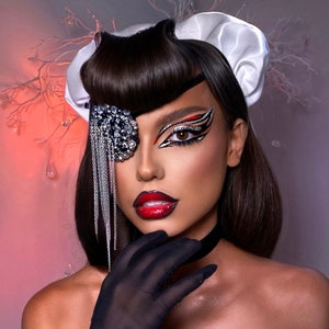 Puede incluir: Una mujer con cabello castaño oscuro y una diadema blanca lleva un maquillaje de ojos dramático con un diseño negro y rojo. También lleva una cadena plateada con un gran adorno de pedrería transparente sobre su ojo izquierdo.