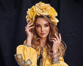 May include: A woman with long blonde hair wears a yellow embroidered blouse and a yellow flower crown. She is posing with her hands near her face.