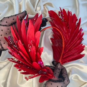 May include: A red feather headband with pink and red jewels. The headband is made of feathers and has a comb on the back for easy attachment.