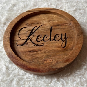 May include: A circular wooden coaster with a raised rim. The coaster is engraved with the name "Keeley" in a cursive font. The wood has a natural grain and a light brown colour.