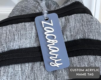 Könnte beinhalten: Ein individuelles Namensschild aus Acryl in rechteckiger Form mit dem Namen "Zachary" in weißer Schrift. Das Schild hat eine gedämpfte blaue Farbe und ist an einem grauen Rucksack mit schwarzen Reißverschlüssen befestigt. Der Text "CUSTOM ACRYLIC NAME TAG" ist ebenfalls sichtbar.