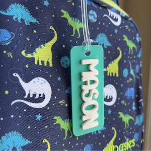 May include: A teal luggage tag with the name "MASON" in white 3D letters hangs in front of a navy blue backpack. The backpack has a dinosaur and space-themed pattern with green, blue, and yellow dinosaurs, planets, and stars.