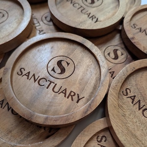 May include: A collection of round wooden coasters with the word "SANCTUARY" and a stylized "S" engraved on each. The coasters are made of light brown wood and have a natural wood grain.