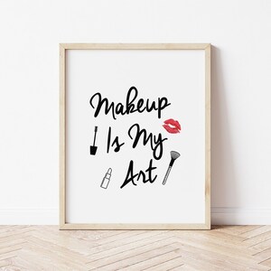 Op de afbeelding: Een ingelijste print met de tekst "Makeup Is My Art" in zwart schrift, vergezeld van illustraties van een mascaraborstel, lippenstift, een make-up kwast en een rode lipafdruk. De lijst is van licht hout.