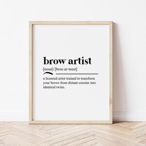 以下が含まれることがあります： 「brow artist」と黒字で書かれた額縁入りのプリント。下の定義は、眉毛を変える訓練を受けたアーティストについて説明しています。フレームは明るい色で、背景は白で、木製の床が見えます。