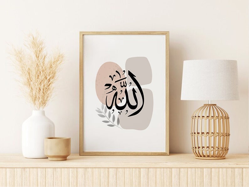 Op de afbeelding: Een ingelijste kunstdruk met zwarte Arabische kalligrafie, tegen abstracte roze en grijze vormen. Het kunstwerk staat op een houten plank, samen met een witte vaas met gedroogd pampasgras, een kleine bruine pot en een decoratieve lamp.