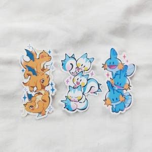 Puede incluir: Tres juegos de pegatinas de dibujos animados con varios personajes. Un juego muestra dragones naranjas con alas azules, otro criaturas azules y blancas con forma de gato, y el &uacute;ltimo criaturas azules con detalles en naranja. Cada juego est&aacute; apilado y rodeado de detalles brillantes.
