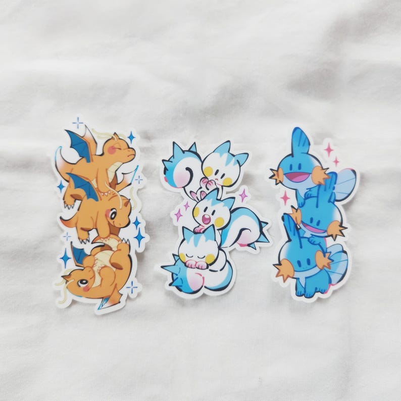 Puede incluir: Tres juegos de pegatinas de dibujos animados con varios personajes. Un juego muestra dragones naranjas con alas azules, otro criaturas azules y blancas con forma de gato, y el &uacute;ltimo criaturas azules con detalles en naranja. Cada juego est&aacute; apilado y rodeado de detalles brillantes.