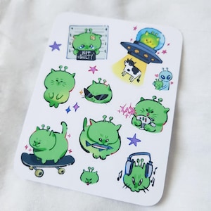 May include: A sticker sheet featuring various green alien cat illustrations. The designs include an alien cat in a spaceship, wearing sunglasses, and holding a fish. Some stickers have text that says "NOT GUILTY".