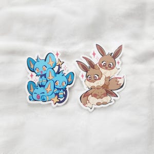 Puede incluir: Dos pegatinas con personajes de dibujos animados. Una pegatina muestra tres criaturas azules con detalles de estrellas. La otra pegatina muestra tres criaturas marrones con detalles esponjosos. Ambas pegatinas tienen un borde blanco y un efecto brillante.