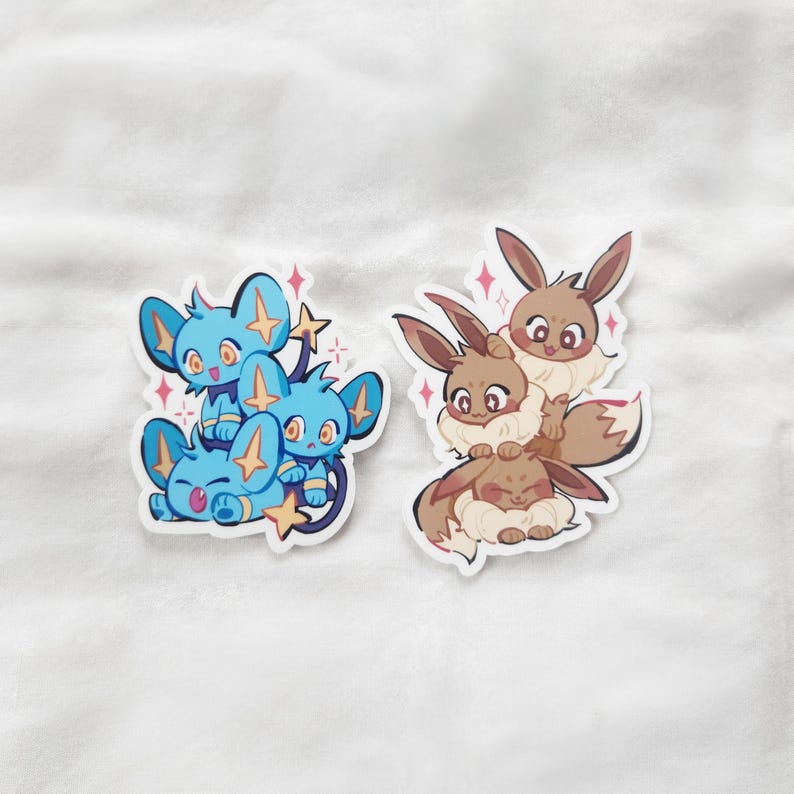 Puede incluir: Dos pegatinas con personajes de dibujos animados. Una pegatina muestra tres criaturas azules con detalles de estrellas. La otra pegatina muestra tres criaturas marrones con detalles esponjosos. Ambas pegatinas tienen un borde blanco y un efecto brillante.