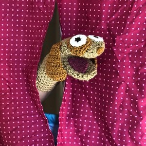 Könnte beinhalten: Eine handgefertigte Häkelpuppe in Form eines Hundes, der hinter einem rosa Polka-Dot-Vorhang hervorlugt. Die Puppe hat Garn in den Farben Beige, Braun und Weiß mit einem lila Mund. Der Vorhang ist leuchtend rosa mit weißen Punkten.