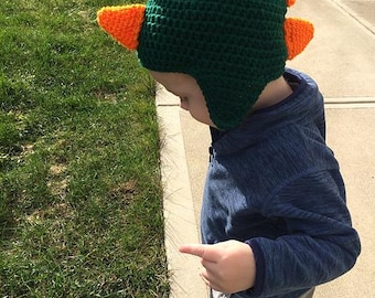 May include: A green and orange crochet hat, resembling a dinosaur, is worn by a child. The hat features a dark green base with orange spikes along the top. The child is wearing a blue hoodie and gray pants.