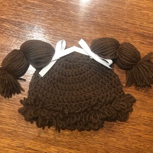 May include: A brown crocheted baby hat with attached yarn pigtails and a white ribbon bow. The hat has a ruffled edge and is designed to resemble a doll's hairstyle. The yarn is a dark brown color.