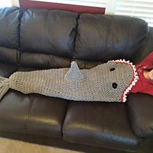 May include: A gray crocheted shark blanket with white teeth and black eyes. The blanket is shaped like a shark with a tail and a fin.