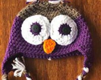 May include: A handmade crochet owl hat in purple, brown, white, and orange. The hat features large white eyes with black button pupils, an orange beak, and ear tufts. The hat has braided ties with tassels.