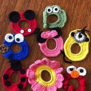 May include: A collection of colorful, crocheted hair ties in various designs. The assortment includes a red Mickey Mouse, a green frog, a blue Cookie Monster, a pink Minnie Mouse, a yellow Minion, a red ladybug, a pink flower, and a red Elmo.