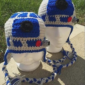 May include: Two handmade crochet hats in a blue, white, and gray color scheme, resembling a popular science fiction character. The hats feature a black circular detail, red accents, and braided ties with tassels. The hats are displayed on white mannequin heads.