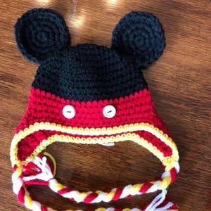 May include: A handmade crochet hat in the style of a cartoon character. The hat is black, red, and yellow, with two large black ears on top. It has white buttons and a braided chin strap. The hat is designed for children.