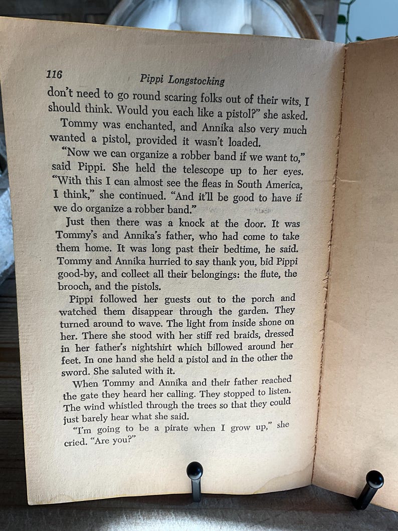 Może przedstawiać: Otwarta strona książki z tekstem z "Pippi Pończoszanki" Astrid Lindgren. Tekst jest czarnym tuszem na postarzanym, kremowym papierze. Dwie czarne metalowe podp&oacute;rki do książek trzymają strony otwarte.