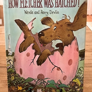 May include: A children's book cover titled "How Fletcher Was Hatched!" features a brown spotted dog emerging from a pink egg. The illustration has a whimsical style with a light blue background and green foliage at the bottom.