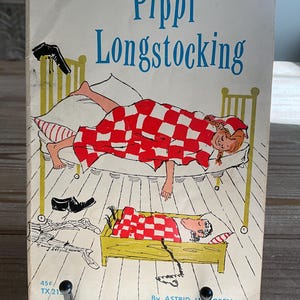 Może przedstawiać: Okładka książki w miękkiej oprawie "Pippi Pończoszanka" autorstwa Astrid Lindgren. Okładka przedstawia ilustrację Pippi w ł&oacute;żku pod czerwoną i białą kratkowaną kołdrą. Tytuł jest w kolorze niebieskim.