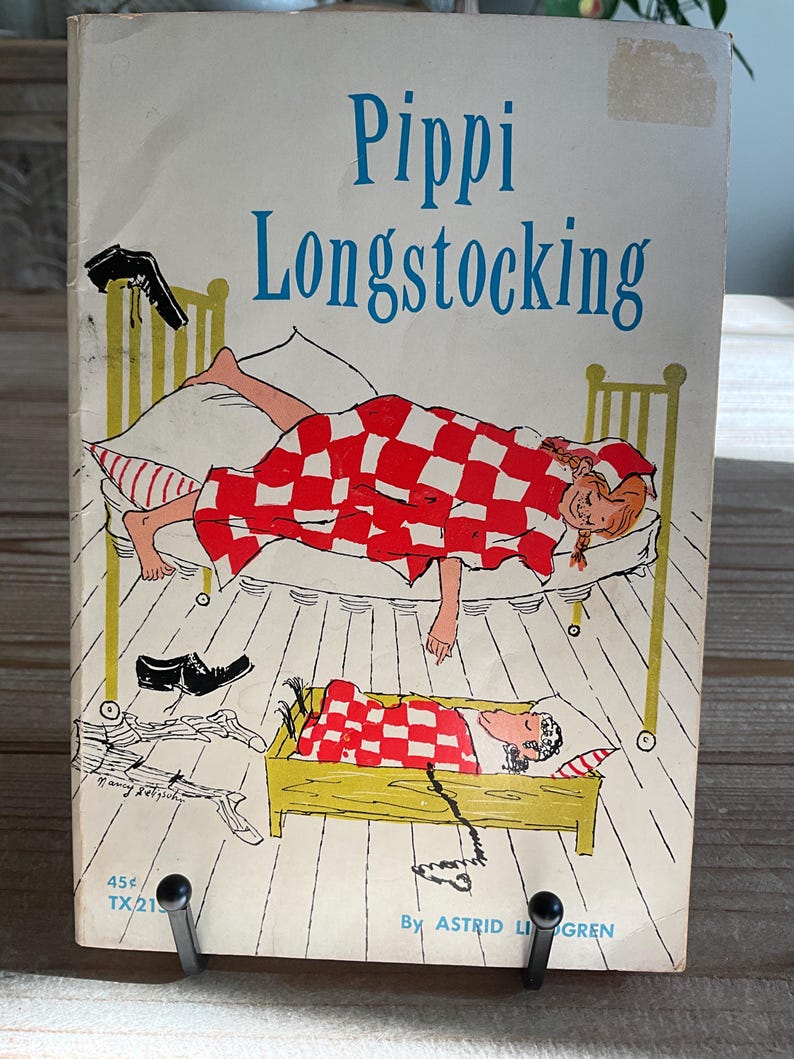 Może przedstawiać: Okładka książki w miękkiej oprawie "Pippi Pończoszanka" autorstwa Astrid Lindgren. Okładka przedstawia ilustrację Pippi w ł&oacute;żku pod czerwoną i białą kratkowaną kołdrą. Tytuł jest w kolorze niebieskim.