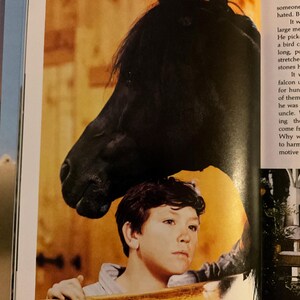 May include: A black horse's head looms over a young person, both positioned behind a wooden fence. The horse's dark coat contrasts with the person's light skin and the warm tones of the wooden structure.