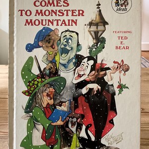 May include: A vintage children's book titled "Christmas Comes to Monster Mountain" with illustrations of monsters and a witch. The cover features the text in red and the book is held by a black metal stand. The book is from the "Good Friends Ideals" series.