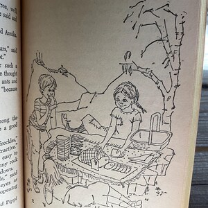 Może przedstawiać: Czarno-biała ilustracja z książki dla dzieci przedstawiająca scenę piknikową. Dwoje dzieci siedzi w pobliżu kosza piknikowego i jedzenia, a trzecie dziecko leży. W tle drzewo i g&oacute;ra.