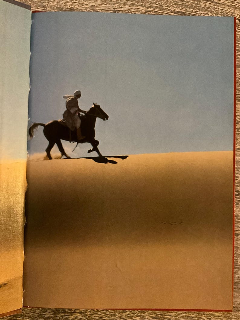 May include: A person riding a dark brown horse across a sand dune under a clear blue sky. The horse is in motion, kicking up sand. The rider is wearing light-coloured clothing and a head covering.