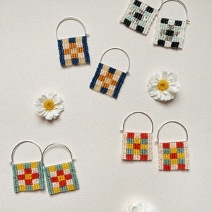 May include: A set of six pairs of geometric patterned hoop earrings. The earrings are made with colorful beads and silver hoops. The patterns include squares, checks, and stripes in shades of blue, green, yellow, red, and white.