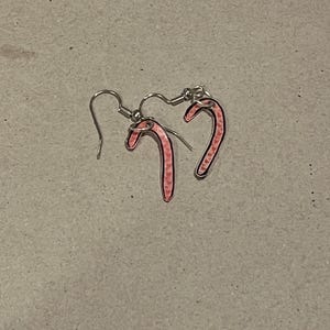 May include: A pair of candy cane earrings. Each earring features a pink candy cane shape with a black outline and a silver-colored hook. The earrings are set against a neutral background.