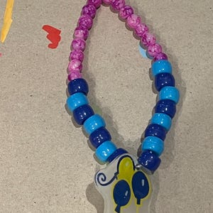 May include: A beaded necklace with a pendant. The necklace features alternating dark blue and light blue beads, transitioning to pink and purple beads near the top. The pendant is a translucent shape with a yellow top and blue balloon design.