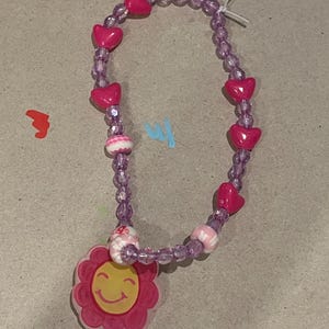 May include: A pink beaded necklace with heart-shaped beads and a flower pendant with a smiling face. The necklace is strung on a purple cord and has a white string closure.