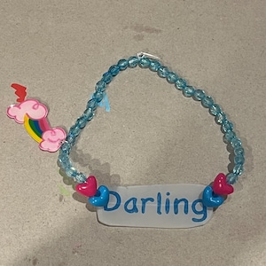 May include: A blue beaded bracelet with the word "Darling" in blue lettering on a white rectangular charm. The bracelet has pink and blue heart-shaped accents and a rainbow charm.
