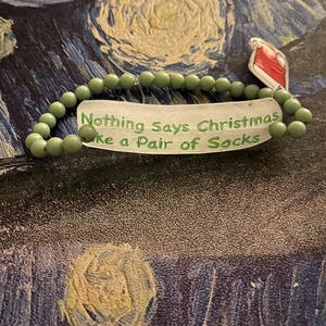 May include: A beaded bracelet with olive green beads and a tag that reads "Nothing Says Christmas Like a Pair of Socks." The bracelet is displayed on a textured background with a painting-like appearance.