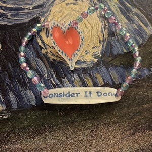 May include: A beaded ornament with a red heart and the words "Consider It Done." The ornament features a string of blue and purple beads. The heart has a white border with blue accents.