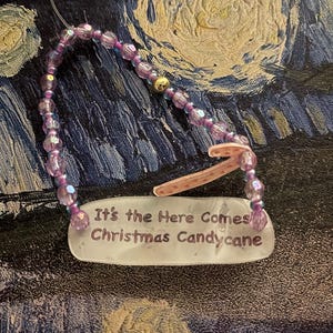 May include: A Christmas ornament with a clear rectangular tag that reads "It's the Here Comes Christmas Candycane." The ornament has a pink candy cane and is strung with purple beads and a gold bead.