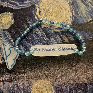May include: A beaded bracelet with blue and clear beads. A white, oval-shaped charm with the text "So Many Clouds" in blue script is attached. A cloud-shaped charm is also attached to the bracelet.