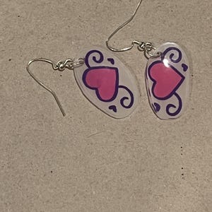May include: A pair of dangle earrings featuring a pink heart design with purple swirls on a clear, oval-shaped background. The earrings have silver-colored hooks.