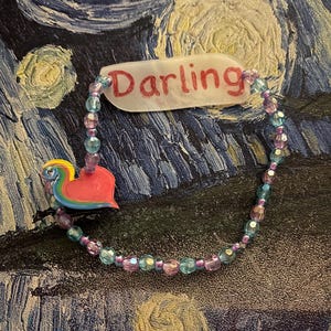 May include: A beaded bracelet with the word "Darling" on a white tag. The bracelet features a mix of pink, blue, and purple beads. A colorful heart-shaped charm is attached to the bracelet. The background is a textured, dark blue and yellow painting.