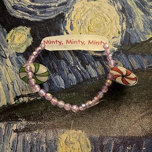 May include: A beaded bracelet with a light purple hue, featuring two charms: one green and white peppermint candy and one red and white peppermint candy. The bracelet has the text "Minty, Minty, Minty" on a white tag.