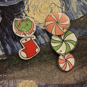 May include: A pair of dangling earrings with a holiday theme. One earring features a green cartoon pony and a red stocking. The other earring has three circular charms with pink and green spiral designs. The earrings have silver-colored hooks.