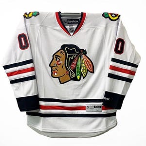 May include: White hockey jersey with red, black and white stripes on the sleeves. The jersey features a large logo of a Native American head with a feather headdress and the words "Reebok" on the front. The number "0" is on both the left and right sleeves.